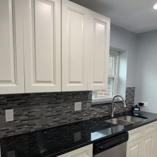 White kitchen cabinets above a black countertop with a silver faucet and sink, gray backsplash.