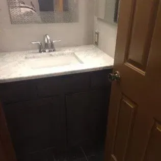 Dark brown vanity with a white countertop and silver faucet in a bathroom. Open wooden door on the right.