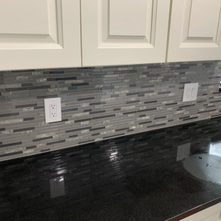 Kitchen with a mosaic tile backsplash and black countertop under white cabinets and outlets.