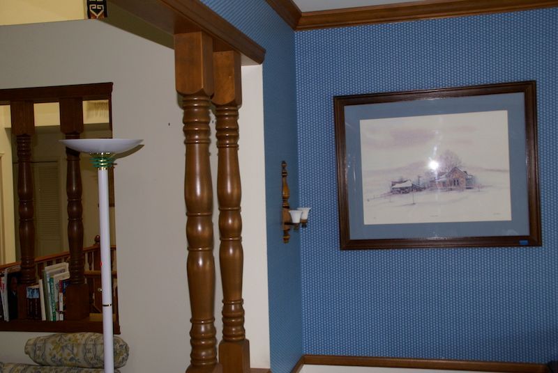 Interior view: blue patterned wall, framed artwork, wooden columns, lamp, mirror.