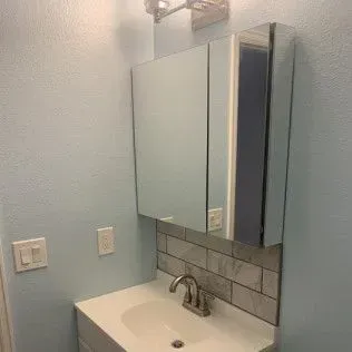 Bathroom with light blue walls, white sink, mirrored medicine cabinet, and tiled backsplash.