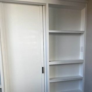 White built-in shelving and sliding door. Shelves are empty. Black door handle. White walls.
