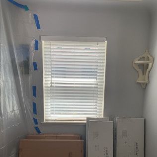 Window with blinds, wall shelf, cardboard boxes, and plastic sheeting taped to the wall.