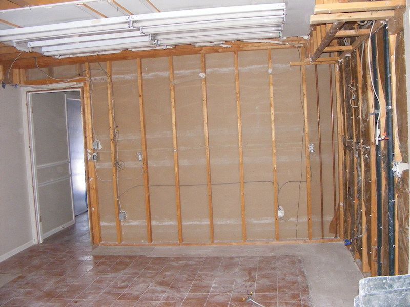 Interior wall demolition exposing wooden studs, electrical wiring, and a doorway in a room with a tiled floor.