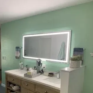 Bathroom with a large, lit mirror above a double sink vanity. The walls are pale green.