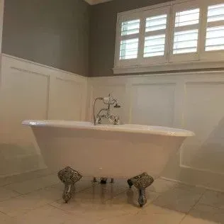 Clawfoot bathtub in a bathroom with white wainscoting and shuttered windows.