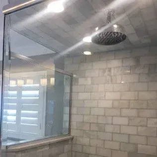 Modern shower with a glass enclosure, white brick-like tiles, a rain showerhead, and a window with white shutters.