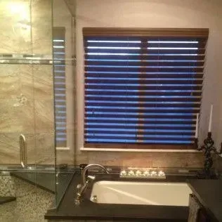 Bathroom with a window covered by blue-toned blinds, a bathtub, and a glass shower enclosure.