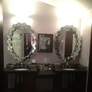 Bathroom with two oval mirrors surrounded by mirrored tiles, black countertops, and dark cabinetry.
