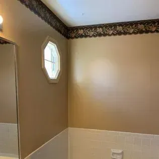 Bathroom corner with tan walls, floral wallpaper border, small window, and a section of mirror.