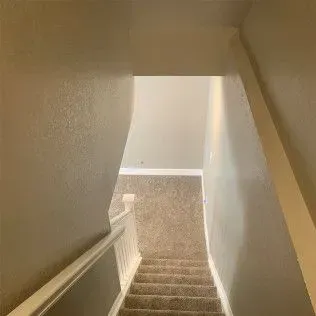 Looking down a carpeted staircase with white railing and walls, leading to a lighter room.
