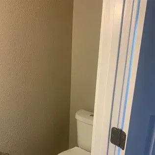 Toilet in a small bathroom with light brown textured walls and a white door with a black handle.