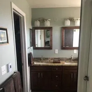 Bathroom with double sinks, mirrors, and cabinetry; door on the left.