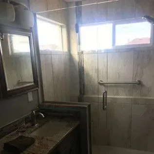 Bathroom with shower, mirror, and sink. Beige tile and glass door. Windows provide natural light.
