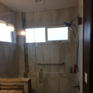 Glass shower with handle and showerhead, light-colored tile walls, and a window.