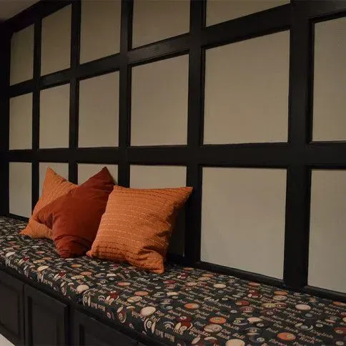 Built-in window seat with patterned cushion, three orange pillows, and grid-paneled wall.