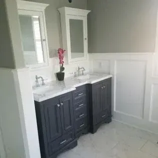 Bathroom with gray cabinets, white countertops, sinks, mirrors, and wainscoting.
