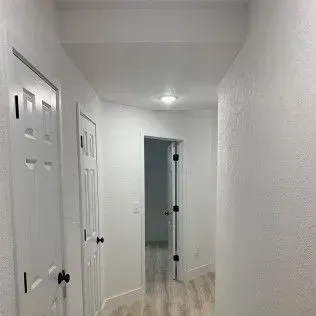 White hallway with doors on the left and center. An open doorway at the end leads to another room.