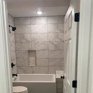 Bathroom with white tub and marble-like tile in the shower. Black fixtures. White door.