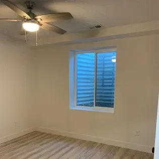 Empty room with a window and a ceiling fan. Pale walls, light wood floor, and blue window view.