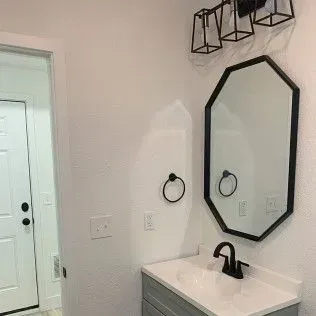 Bathroom with mirror, sink, and lighting fixtures. Black accents on white walls. Doorway to the left.
