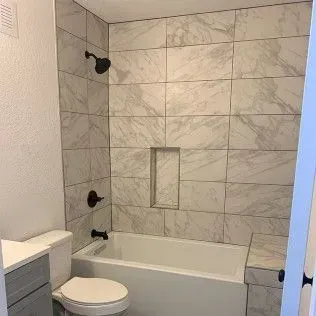 Bathroom with marble-look tiled shower walls, built-in tub, dark fixtures, and a toilet.