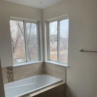 Bathroom with a white bathtub beneath two windows. Tan tile surrounds the tub.