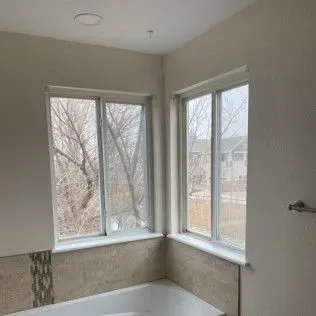 Two white-framed windows above a bathtub, with a view of trees and buildings outside. Beige tile covers the lower walls.