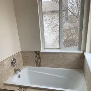 Bathroom with a white bathtub, beige tile, and a window overlooking bare tree branches.