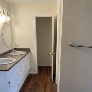 Bathroom with a double sink vanity, an oval mirror, and an open doorway leading to a room.
