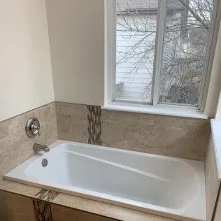 White bathtub in a tiled bathroom with a window overlooking bare trees and a faucet.
