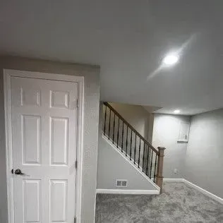 A doorway, staircase, and carpeted room. The walls are gray, with a white door and wood railing.