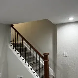 Staircase with brown banister and black spindles, ascending to an upper level. Gray carpeted steps. Beige wall.