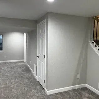 Basement interior with gray walls and carpet. A white door stands near stairs with a black railing.