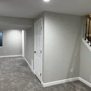 Gray basement with carpet, a white door, and a staircase with black railing.