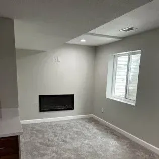 Empty basement room with fireplace, window, gray walls, and carpet.
