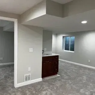 Finished basement with gray carpet, walls, and dark wood cabinetry. A small window is visible.