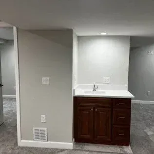 Bathroom vanity in a corner of a room with dark wood cabinets, white countertop, and gray walls and carpet.