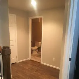 Hallway with hardwood floor, leading to a bathroom and a white door, light beige walls.
