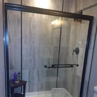 Shower enclosure with black frame and glass doors, gray tiled walls, and a small stool with toiletries.