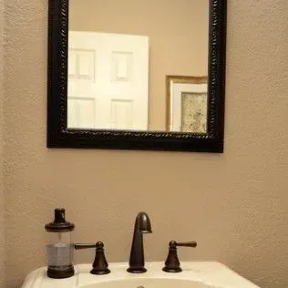 Black-framed mirror above a bathroom sink with bronze fixtures.  Tan walls and white door reflected.