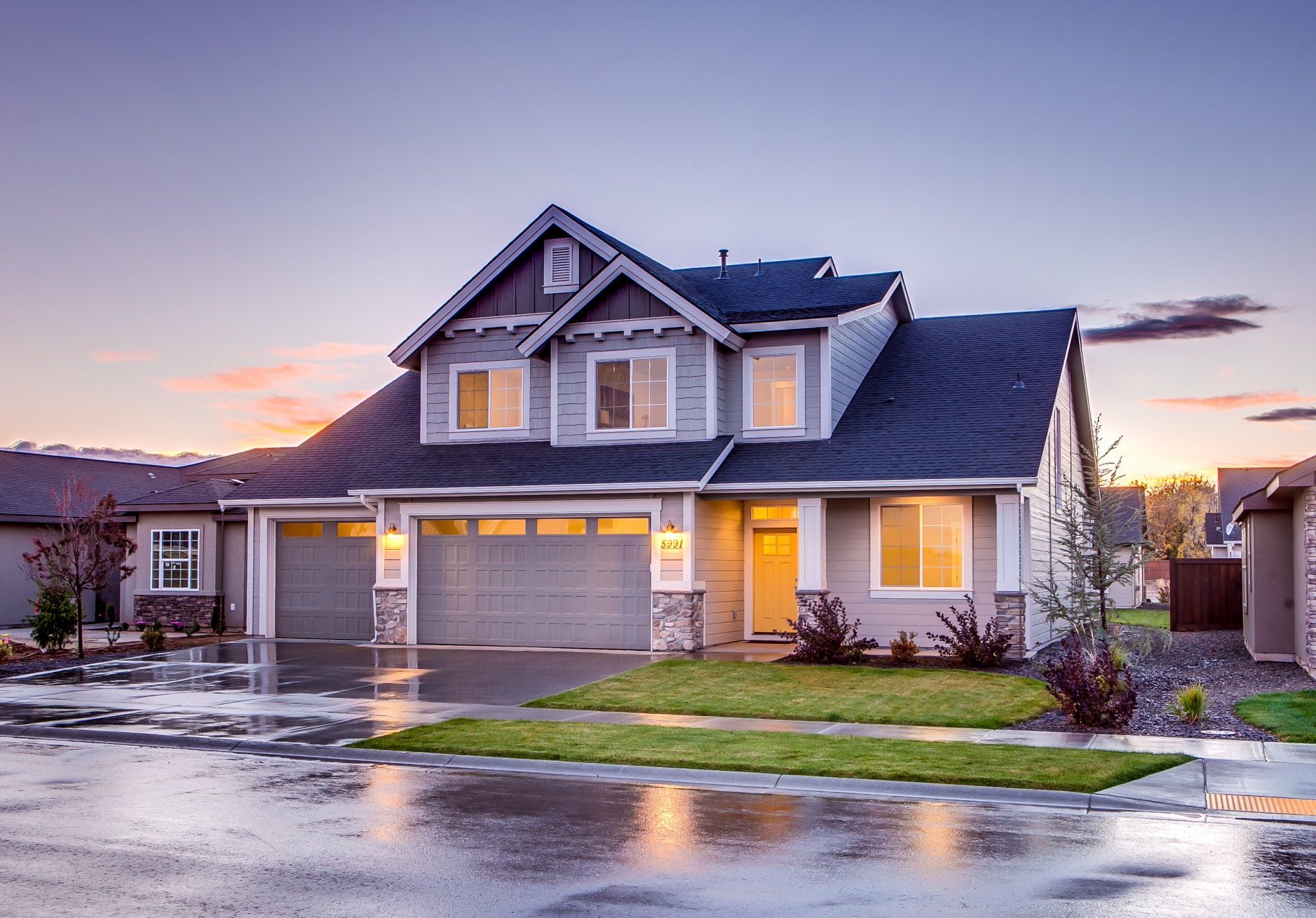 Two-story gray house with a three-car garage, wet driveway, and evening sky.