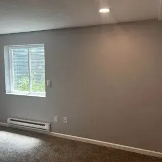 A room with a window, heating unit, and gray walls, with a carpeted floor.