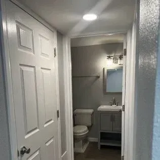Hallway leading to a bathroom; white door on the left, gray bathroom with toilet, sink, mirror.