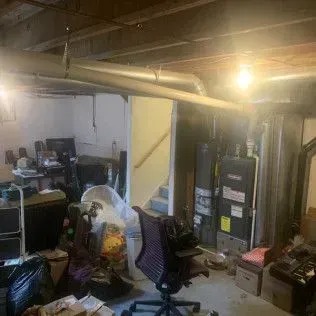 Cluttered basement with ductwork, stairs, and various items like boxes and chairs.