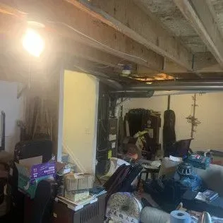 A cluttered basement with exposed beams, piles of items, and a visible light fixture.