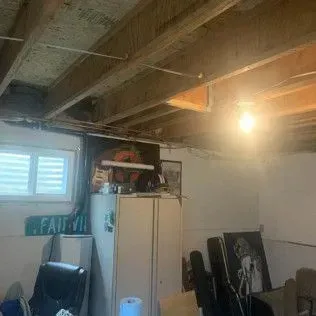 Basement interior with exposed beams, window, appliances, artwork, and various items. Bright light.