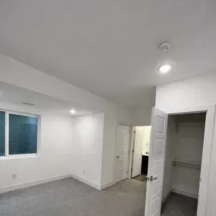 Empty bedroom with closet, window, and recessed lighting. The walls are white and the floor is gray.
