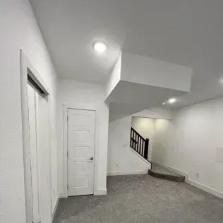 Interior view of a white-walled room with a door, stairs, and recessed lighting. Carpeted floor.