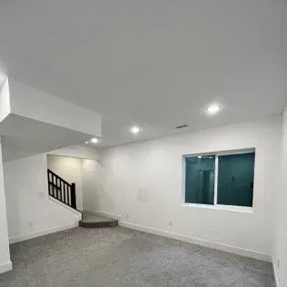 Empty room with gray carpet, a window, stairs leading upwards, and recessed ceiling lights.